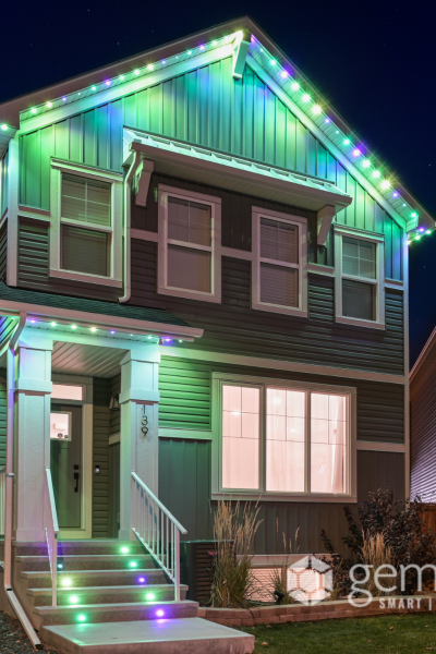 A two-story house with a gable roof is adorned with colorful LED strip lights along its eaves and staircase, creating a vibrant display against the night sky. The house features a covered front porch and a well-manicured lawn, with neighboring houses visible on either side. The Gemstone Lights logo is prominently displayed in the bottom-right corner, highlighting the brand's smart outdoor lighting solutions.