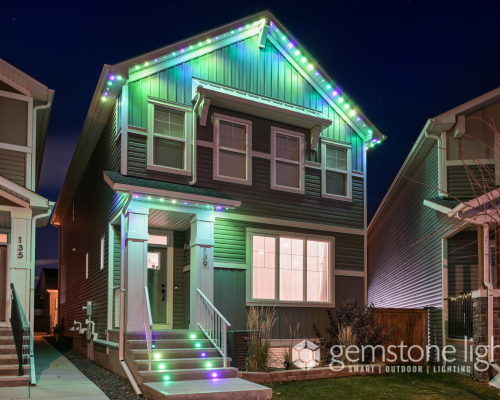 A two-story house with a gable roof is adorned with colorful LED strip lights along its eaves and staircase, creating a vibrant display against the night sky. The house features a covered front porch and a well-manicured lawn, with neighboring houses visible on either side. The Gemstone Lights logo is prominently displayed in the bottom-right corner, highlighting the brand's smart outdoor lighting solutions.