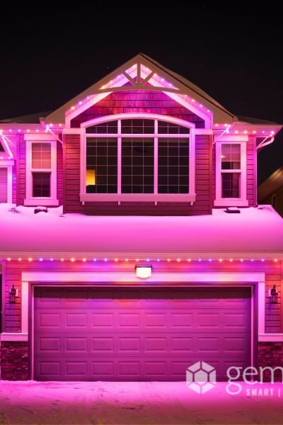 A two-story house adorned with vibrant pink and purple Gemstone Lights, a smart outdoor lighting system, on its roof and garage, standing out in a snowy neighborhood at night.