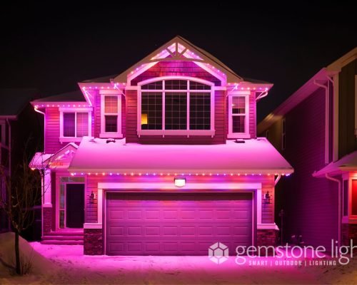 A two-story house adorned with vibrant pink and purple Gemstone Lights, a smart outdoor lighting system, on its roof and garage, standing out in a snowy neighborhood at night.