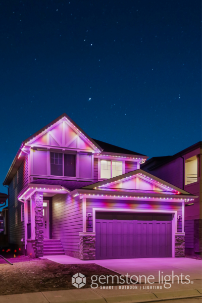 A two-story house with a garage, adorned with vibrant purple and pink LED lights along its roof edges and gutters, set against a dark night sky filled with stars.