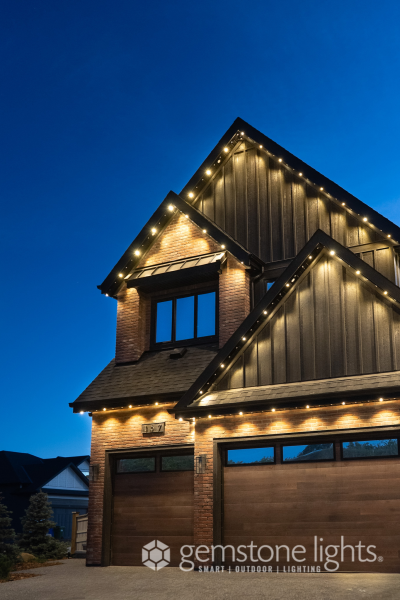 Modern two-story house with brick and dark wood siding, illuminated by warm outdoor string lights along the roofline and eaves, set against a clear blue evening sky.