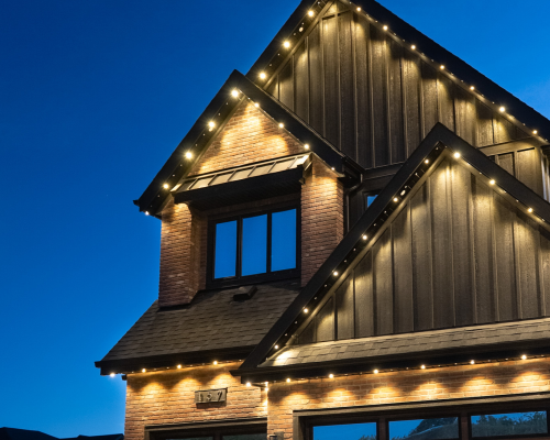 Modern two-story house with brick and dark wood siding, illuminated by warm outdoor string lights along the roofline and eaves, set against a clear blue evening sky.