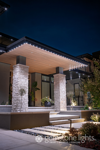 A modern home's front entrance at night, featuring a sleek roof with Gemstone Lights LED strip lighting integrated along the edges, illuminating the pathway and accentuating the stone pillars and contemporary design.