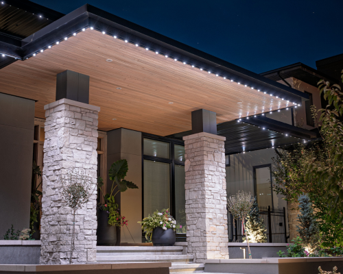A modern home's front entrance at night, featuring a sleek roof with Gemstone Lights LED strip lighting integrated along the edges, illuminating the pathway and accentuating the stone pillars and contemporary design.