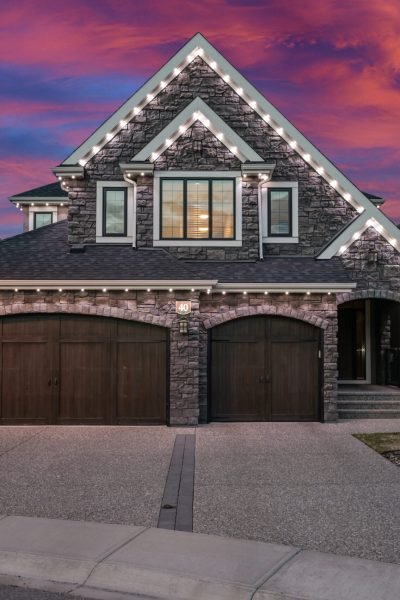 Halo Lighting installation on a modern stone house with a two-car garage, featuring white LED lights along the roof edges and around the garage, set against a vibrant pink and purple sunset sky.