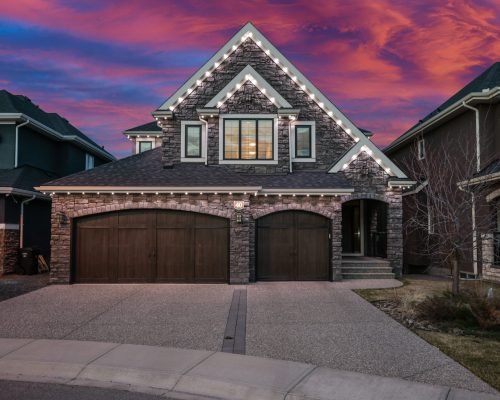 Halo Lighting installation on a modern stone house with a two-car garage, featuring white LED lights along the roof edges and around the garage, set against a vibrant pink and purple sunset sky.
