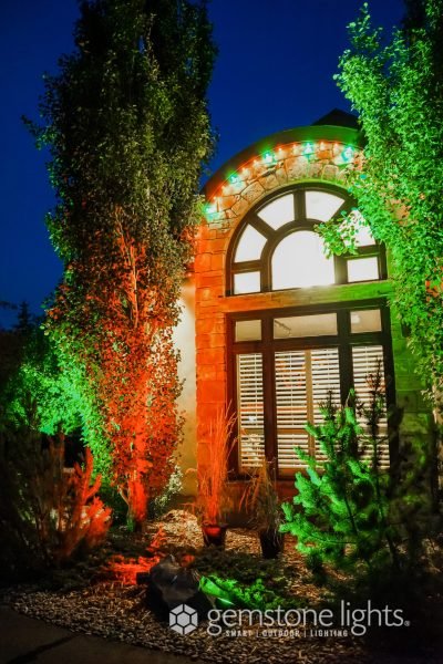 A house facade at night with a large arched window illuminated by green and orange uplighting, complemented by colorful accent lights along the archway and bright white light inside the window, surrounded by lush greenery.