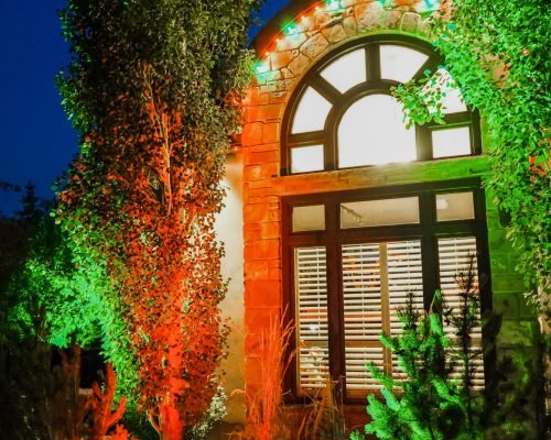 A house facade at night with a large arched window illuminated by green and orange uplighting, complemented by colorful accent lights along the archway and bright white light inside the window, surrounded by lush greenery.